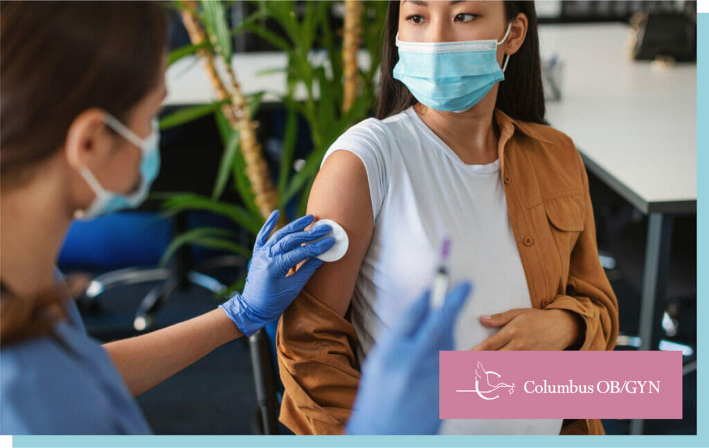 Female doctor in protective face mask and gloves preparing to inject pregnant patient with covid antiviral vaccine, rubbing hand with cotton pad