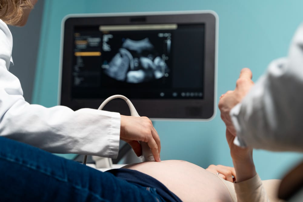 OB-GYN provider performing an ultrasound exam on a pregnant patient, with fetal imaging displayed on a medical monitor.